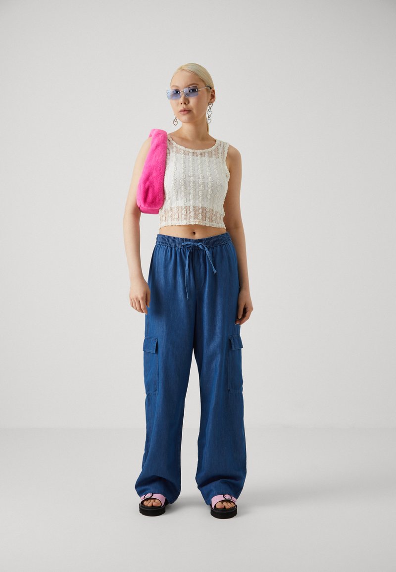 Top court en dentelle, pantalon cargo large bleu avec poches et sac en fausse fourrure rose. Des sandales noires avec des accents roses complètent le look.