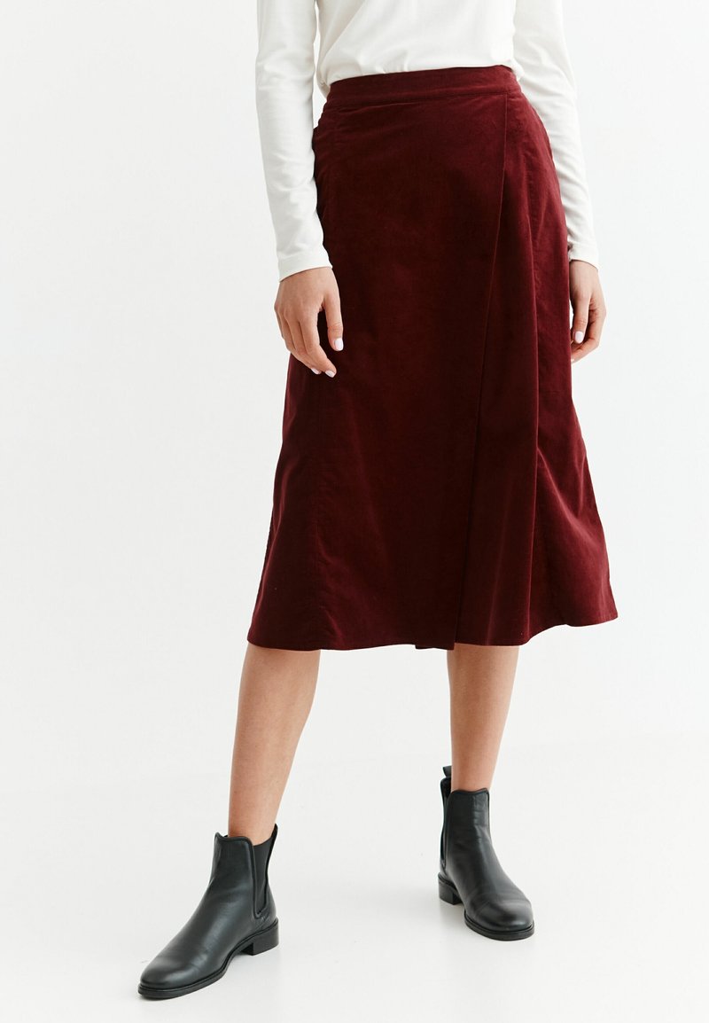 Femme portant une jupe midi bordeaux, un haut blanc à manches longues et des bottines noires, debout devant un fond blanc uni.