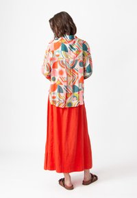 Kleurrijke blouse met abstract patroon en korte mouwen, gecombineerd met een lange, vloeiende oranje rok. Bruine sandalen zichtbaar bij de voeten.