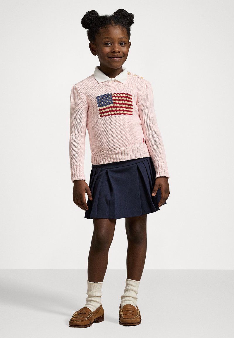 Suéter rosa tricotado com um padrão da bandeira dos EUA, combinado com uma saia plissada azul-marinho. Mocassins bege e meias creme completam o conjunto.