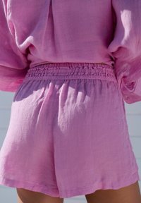 Kvinna klädd i rosa linnebyxor och matchande långärmad topp, sedd från midjan neråt och bakifrån i naturligt ljus.