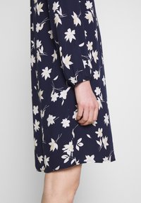 Robe bleu marine à motif floral blanc, manches longues resserrées aux poignets, montrée de la moitié de la cuisse à l'avant-bras sur une personne à la peau claire.