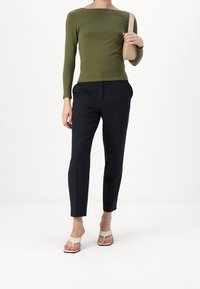 Olivgrön figurnära långärmad topp, kombinerad med skräddarsydda mörkblå byxor. Modellen bär beige sandaler och håller en liten beige väska.