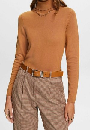 Kobieta w kremowym swetrze z golfem, z łańcuszkowym naszyjnikiem, brązowych plisowanych, w kratę spodni oraz beżowym skórzanym pasku z srebrną klamrą.