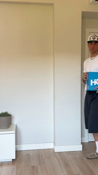 Caixa azul com logo branco segurada nas mãos, colocada numa sala minimalista com piso de madeira clara e um vaso cinza contendo uma planta.