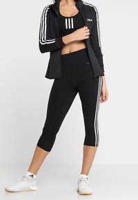 Černý sportovní outfit se zipovou bundou, krátkým topem a capri legínami. Má bílé pruhy na rukávech a bocích. Materiál vypadá pružně.