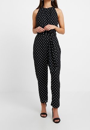 Kvinde iført sort ærmeløs halterneck jumpsuit med hvide polkaprikker og matchende stofbælte, kombineret med sorte åbne hæle.