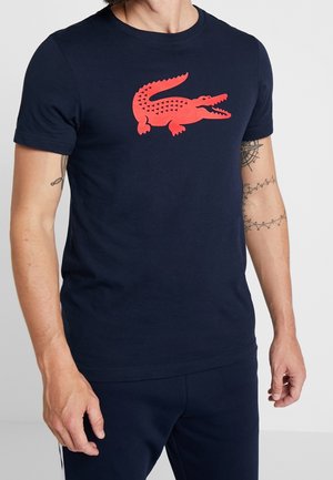 Mężczyzna w granatowej koszulce z dużym czerwonym logo krokodyla na piersi i pasujących granatowych spodniach, prezentujący tatuaże na ramionach.