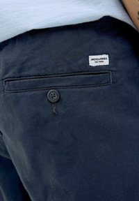 Shorts en coton navy avec une poche arrière dotée d'un bouton noir et d'une étiquette portant l'inscription "JACK&JONES EST.1990." La texture semble lisse.