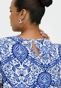 Kvinna med mörkt hår i en knut som bär silverfärgade hoops och en vit blus med intrikata blå blommönster, handen vilande på sin nacke.