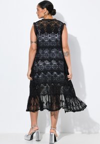 Svart spetsklänning med intrikata blommönster, ärmlös med en volangkant, med en genomskinlig textur och design på ryggen. Silverfärgade klacksandaler.