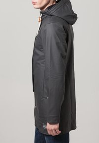 Veste grise imperméable avec capuche, fermeture éclair sur le devant et poches latérales. Texture lisse avec un design relaxé et à coupe droite.