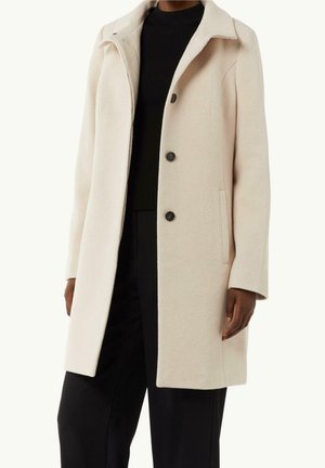 Personne portant un manteau en laine beige arrivant aux genoux avec trois boutons, sur un pull noir et un pantalon noir, se tenant devant un fond neutre.