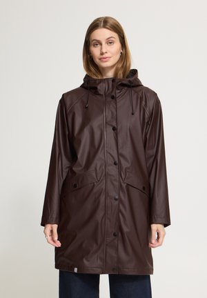 Femme portant un long imperméable marron foncé à capuche avec boutons-pression et poches à rabat, debout devant un fond clair uni.