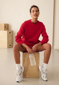 Röd sweatshirt med ribbade muddar, sittande på en trälåda, håller en genomskinlig vattenflaska, bär vita strumpor och grå sneakers.