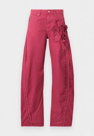 Femme debout, les mains dans les poches, portant un haut blanc à manches courtes, un pantalon large rose avec broderie florale et des chaussures noires ouvertes au bout.