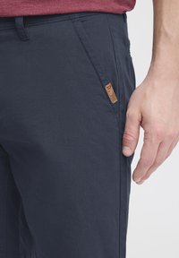 Navy blue pants with a textured fabric, featuring a subtle logo patch on the side pocket and a straight cut design.