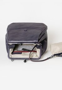 Zaino in pelle di un blu scuro con chiusura a ribalta e bottone, dotato di un interno a righe e una tasca zip rossa. Piccola pochette rimovibile nelle vicinanze.