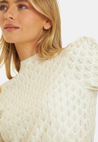 Jasno beżowa bluzka z teksturowanym wzorem w diamentowe kształty i metalicznymi akcentami. Krótkie bufiaste rękawy i wysoki dekolt.