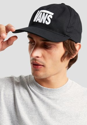 Giovane uomo che indossa un cappellino Vans nero, che regola la visiera con la mano destra, vestito con una maglia girocollo grigio chiaro su uno sfondo semplice.