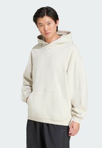 Felpa color crema con tasca a marsupio, texture morbida e un piccolo logo tonale sul petto. Abbinata a pantaloni neri larghi.