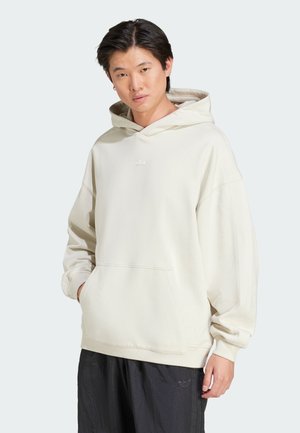 Haut à capuche crème avec une poche kangourou, texture douce, et un petit logo ton sur ton sur la poitrine. Associé à un pantalon noir ample.