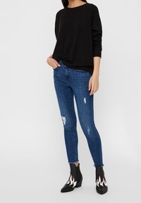 Top negro de manga larga, jeans ajustados azules con detalles desgastados y botas negras hasta el tobillo con acentos de llamas blancas.