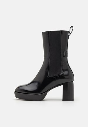 Bottine en cuir verni noir avec talon carré, panneau latéral élastique et languette de tirage sur fond blanc.