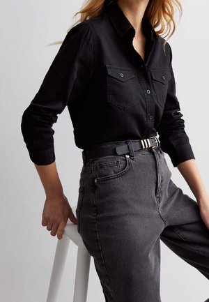 Vrouw in een zwart overhemd met knopen, gestopt in een hoog getailleerde donkergrijze spijkerbroek, zittend op een witte kruk, hand rustend op de achterkant van de kruk.