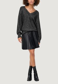 Maglione nero a maglia con scollatura a V e maniche lunghe, abbinato a una gonna plissettata nera e stivaletti neri alla caviglia. Outfit casual su sfondo neutro.