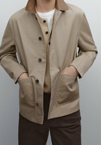 Beige lichtgewicht jack met bruine kraag, knoopsluiting en twee voorzakken. Gedragen over een beige gebreide top en donkere broek. Eenvoudig ontwerp.