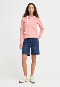 Rožinis užsegamas džemperis su baltais raišteliais, priekinėmis kišenėmis ir džinsinės šortai, siekiantys kelius. Modelis dėvi smėlio spalvos sportinius batelius, demonstruojančius kasdieninį stilių.