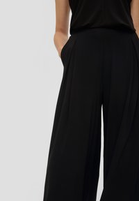 Zwarte wijde pantalons met een soepele textuur, voorzien van zijzakken en subtiele plooien bij de taille voor extra dimensie.