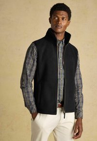 Melnā vilnas veste ar augstu apkakli virs daudzkrāsaina urķa krekla. Iespējas sānu kabatām un priekšējam rāvējslēdzējam. Krēmbalti bikses.