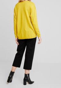 Gele oversized gebreide trui met lange mouwen, gecombineerd met zwarte wijde broek en zwarte enkelboots met een ritsdetail.