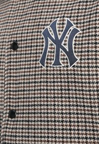 Jachetă în carouri cu un model în culorile negru, alb și maro, având un logo brodat NY de culoare bleumarin și detalii cu nasturi negri.