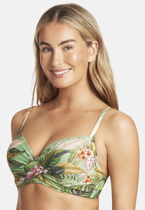 Uśmiechnięta blondynka w zielonym kwiatowym topie bikini z cienkimi ramiączkami na jednolitym białym tle.