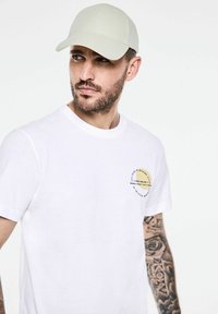 Bílý krátký rukáv tričko s kulatým grafickým logem v žluté a černé barvě, nošeno se světle zelenou kšiltovkou. Viditelný tetování na předloktí.