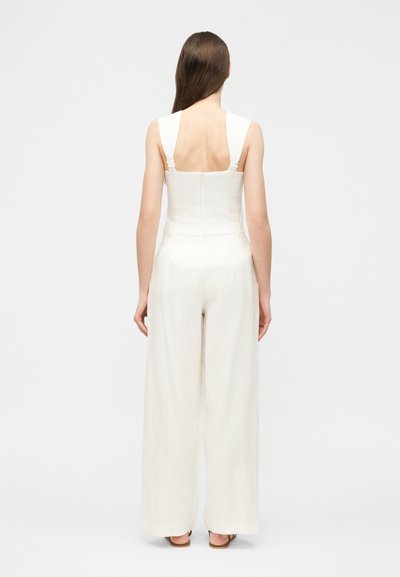 Γυναίκα με μακριά καστανά μαλλιά που φοράει άσπρη ολόσωμη φόρμα χωρίς μανίκια με φαρδιά μπατζάκια, στέκεται γυρισμένη προς τα πίσω σε απλό φόντο.