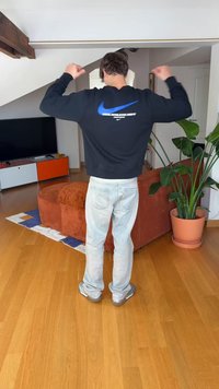 Sweatshirt preto da Nike com logótipo azul, jeans azul claro e ténis cinzentos; chão de madeira e planta em vaso no interior ao fundo.