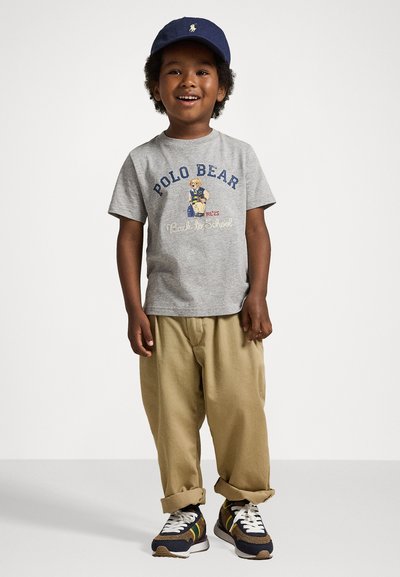 Grå t-skjorte med "Polo Bear" trykk, beige bukser og svarte joggesko med gule detaljer. Marineblå cap fullfører antrekket.