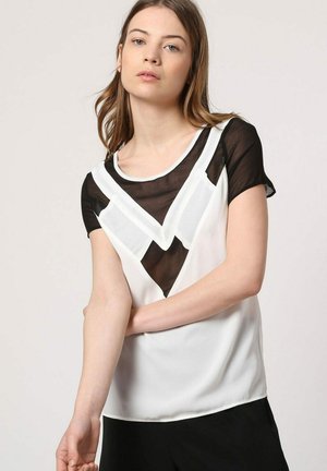 Mujer con blusa de manga corta en blanco y negro con paneles geométricos translúcidos, posando con un brazo doblado sobre la cintura.