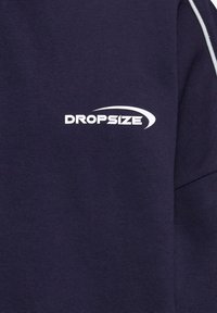 Vêtement bleu marine comportant le texte blanc « DROPSIZE » avec un motif en virgule incurvée au-dessus de la zone droite de la poitrine.