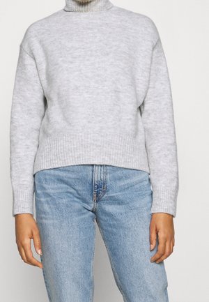 Persoon die een lichtgrijze geribbelde coltrui en hooggeleegde lichtblauwe spijkerbroek draagt, staand met ontspannen armen langs het lichaam.