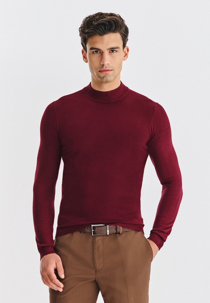 Bordowy dzianinowy sweter z golfem, z długimi rękawami, o dopasowanym kroju i ściągaczami na mankietach. Noszony z brązowymi spodniami i brązowym paskiem.