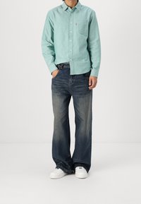 Persona che indossa una camicia a maniche lunghe verde chiaro con bottoni, jeans larghi blu sbiaditi e sneakers bianche, in piedi contro uno sfondo neutro.
