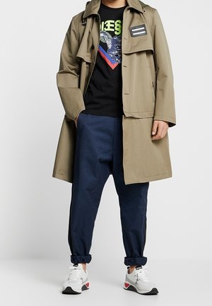 Gabardina color caqui sobre una camiseta gráfica negra, combinada con pantalones azul marino sueltos y zapatillas blancas con detalles rojos. Cuello plano, gran bolsillo delantero.