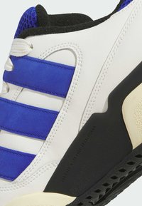 White sneaker with blue suede stripes, black rubber sole, and textured upper. Features stitching details and design markings on the sole.