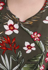 Wydrukowana tkanina o ciemnozielonym tle ma duże białe i czerwone wzory kwiatowe oraz liściaste akcenty. Widoczny detal dekoltu w kształcie litery V.