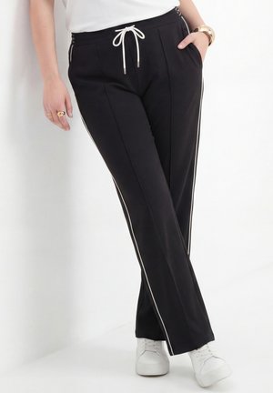 Persoon draagt zwarte broek met witte zijstrepen en een witte aantrekkoord, gecombineerd met witte sneakers, staand tegen een witte muur.